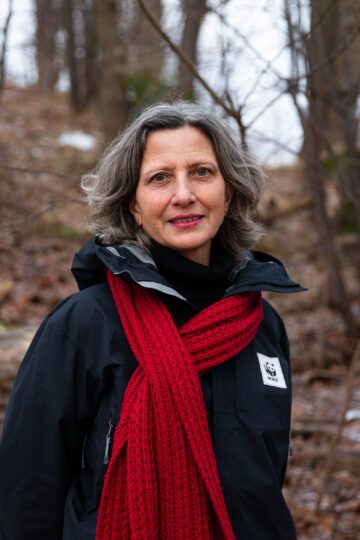 Porträttfoto av Maria Schultz med grått hår iförd svart jacka med WWF-logga och röd halsduk i en skogsmiljö.