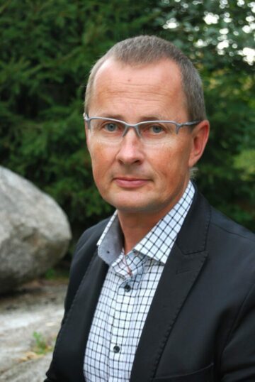 Porträttfoto av Magnus Emfel iklädd mörk kavaj och rutigt skjorta står utomhus, med gröna buskar och en stor sten i bakgrunden.