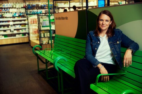 Monika Magnusson sitter på en grön bänk inomhus i en butiksmiljö, omgiven av hyllor med varor och kylskåp i bakgrunden. Hon har på sig en jeansjacka och ett avslappnat uttryck.