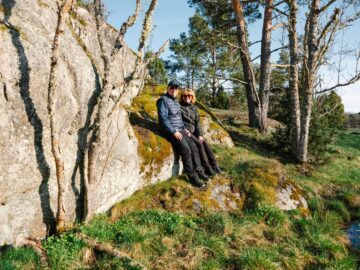 Två personer sitter tillsammans på en mossig stenbumling omgivna av träd och grönska under en solig dag.