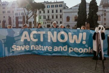En grupp människor håller upp en blå banderoll med texten: "ACT NOW! Save nature and people!" framför byggnader och träd i stadsmiljö.