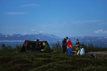En grupp människor sitter och står vid ett tält på en gräsbevuxen kulle med snötäckta berg och en klar blå himmel i bakgrunden.