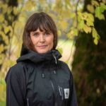 elin-larsson Porträttfoto av Elin Larsson, med mörkbrunt hår och svart jacka och står utomhus framför träd med gulgröna löv.