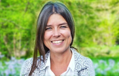 kristinaatkisson Porträttfoto av Kristina Atkisson med långt mörkt hår ler mot kameran. Hon har en vit skjorta och en mönstrad jacka, med en grön bakgrund av träd och natur.