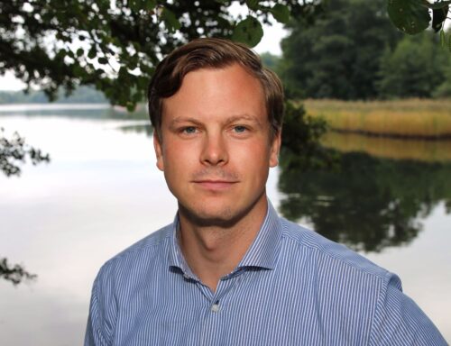 Porträttfoto av Martin Wadmark, med kort hår och skjorta som står framför en sjö omgiven av träd och vass.