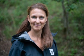 Porträttfoto av Moa Engström med långt brunt hår ler och bär en mörk jacka med WWF-logotypen. Bakgrunden är en skogsmiljö.