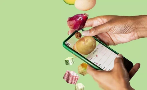 En hand håller en grön smartphone som visar en shoppinglista, medan olika matvaror som potatis, lök, smör och ägg svävar nedåt en grön bakgrund.