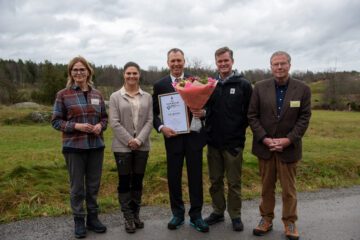 En grupp på fem personer står utomhus på en mulen dag, en av de är kronprinsessan Victoria, WWFs generalsekreterare Gustaf Lind. Erik Ahlström står i mitten och håller en bukett blommor och ett diplom.