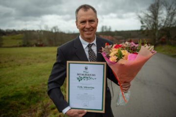 Porträttfoto av Erik Ahlström i kostym som håller en inramad utmärkelse och en bukett blommor; han står utomhus med en grönskande landsbygd i bakgrunden.