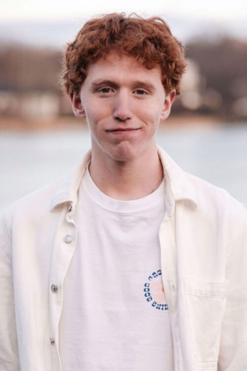 Porträttfoto av Alexander Engströmer med rött lockigt hår står utomhus och bär en vit skjorta över en vit t-shirt med ett tryckt mönster. Bakgrunden är suddig med vatten och träd.