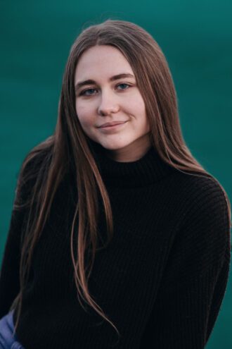 disa Porträttfoto av Disa Ljungman med långt brunt hår och en svart tröja står framför en grön bakgrund och ler lätt.