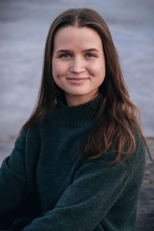 Porträttfoto av Emma Hellström med långt, brunt hår och en mörkgrön tröja ler mjukt mot kameran, med en suddig bakgrund med vatten.