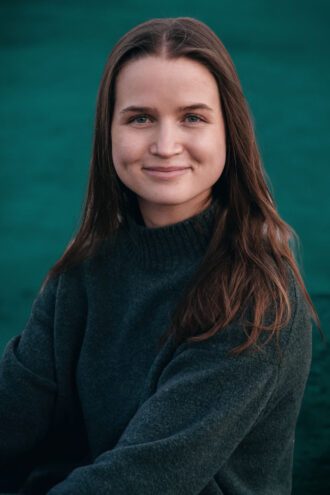 emma Porträttfoto av Emma Hellström med långt, mörkt hår och en mörkgrön tröja ler vänligt och tittar in i kameran. Bakgrunden är suddig och grön.