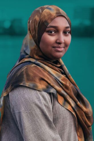 farhiyo-1 Porträttfoto av Farhiyo Muse klädd i en mönstrad sjal och grå klädsel står utomhus framför en suddig, grön bakgrund och tittar mot kameran med ett mjukt leende.