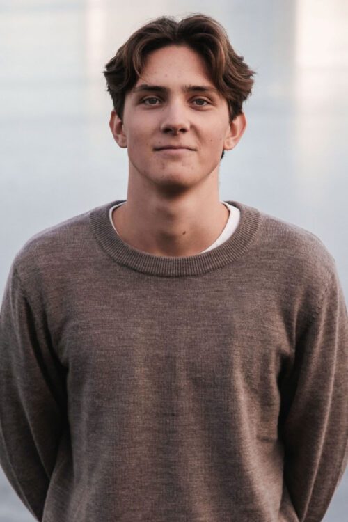 Porträttfoto av Holger Ehrström med kort mörkt hår bär en brun tröja och står framför en suddig bakgrund med vatten.