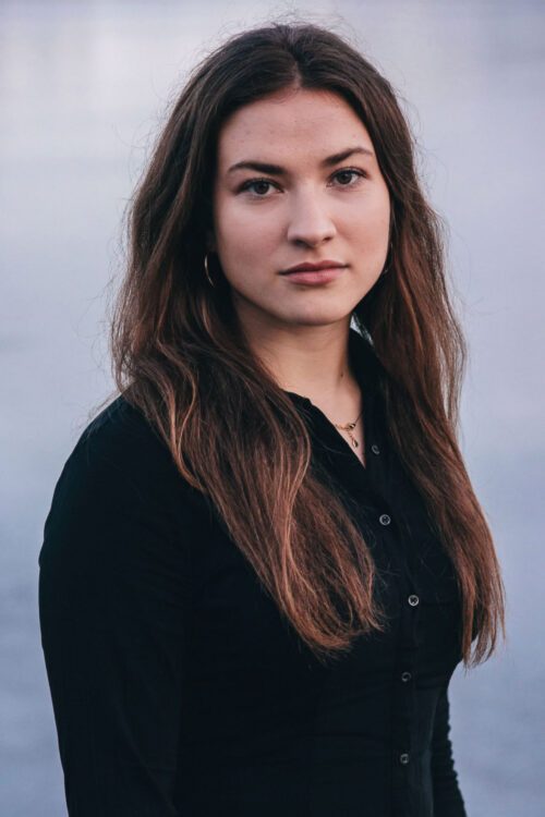 Porträttfoto av Michelle Marinkovic med långt, brunt hår bär en svart skjorta och står utomhus med en suddig, grå bakgrund med vatten.