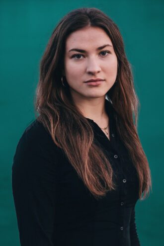 michelle Porträttfoto av Michelle Marinkovic med långt brunt hår och svart skjorta står framför en enfärgad grön bakgrund.