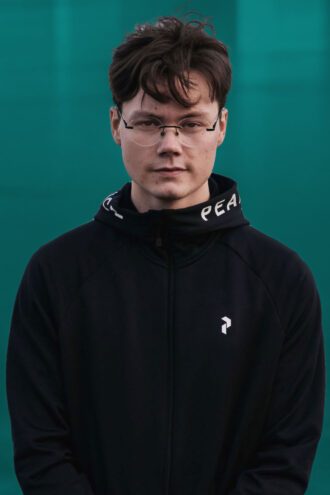 nils-gustu Porträttfoto av Nils-Gustu Kuhmunen med glasögon och mörkt hår bär en svart tröja med texten "PEACE" på kragen, mot en grön bakgrund.