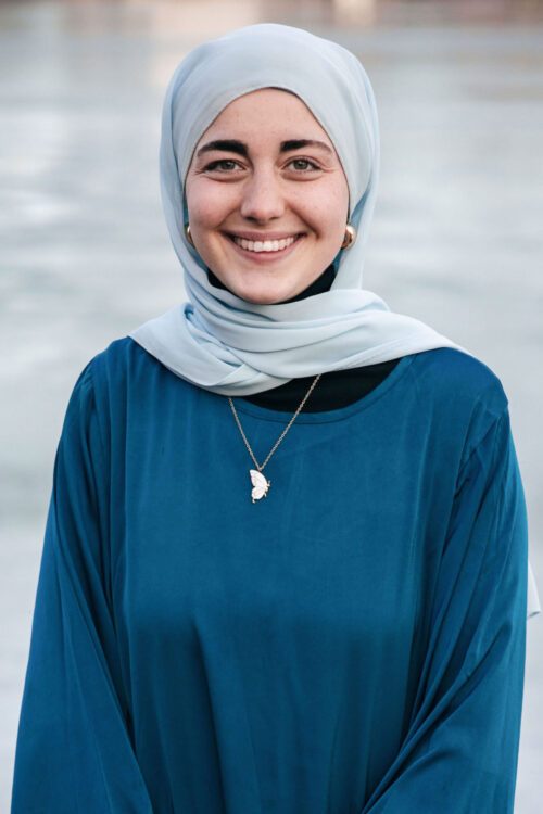 Porträttfoto av Shaden El Seedi bär en ljusblå hijab och en mörkblå tröja. Hon har ett halsband med en måneformad berlock och står framför en suddig bakgrund med vatten.