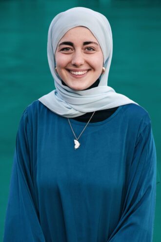 shaden Porträttfoto av Shaden El Seedi med ljusblå hijab och blå klänning står framför en grön bakgrund och bär ett halsband med ett fjärilhänge.