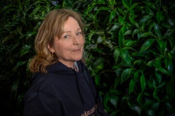 Porträttfoto av Marianne Nordin, med mörk blont hår bär en mörkblå tröja och står framför en bakgrund av frodiga gröna växter.