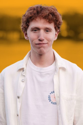 Porträttfoto av Alexander Engströmer med lockigt rött hår bär en vit skjorta över en vit t-shirt med en liten cirkellogga. Bakgrunden är suddig med gula och gröna toner.