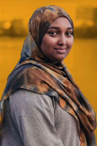 Porträttfoto av Farhiyo Muse i en brun och beige hijab står utomhus och tittar mot kameran, med en suddig gul bakgrund.