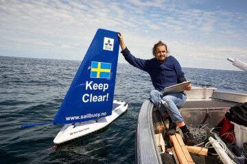 En person sitter i en båt på havet och håller i en modellsegelbåt med en blå segel som har en svensk flagga, texten "Keep Clear!" och webbadressen "www.sailbuzzy.com" syns på modellsegelbåtens segel.