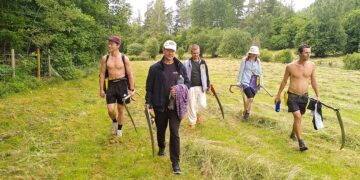 Fem personer går över en grön äng, hållandes liar och redskap. Skogen syns i bakgrunden.