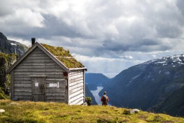 En liten trästuga med grästak står på en grön kulle med en person som vandrar nära kanten, där en djup fjord och berg syns i bakgrunden under molnig himmel.