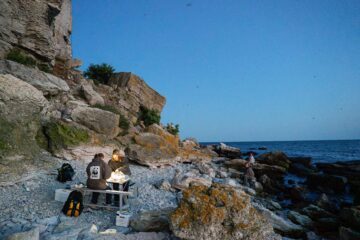Två personer sitter vid ett campingbord på en stenig strand nära havet, med klippor och växter i bakgrunden under en klar blå himmel.