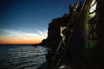 Personer klättrar uppför en stege vid en klippa nära havet, med en färgstark solnedgång i bakgrunden.