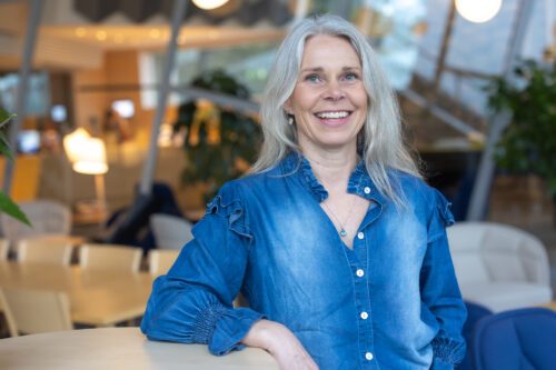 Porträttfoto av Annica Gilbertson, ljust hår och jeansblå skjorta lutar sig mot ett ståbord. Ler mot kameran.