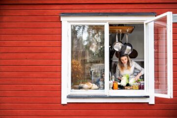 Anastasia Lundqvist står vid ett öppet köksfönster, husets fasad är rött med vitt fönsterbräde. Anastasia håller i olika grönsaker inne i köket.