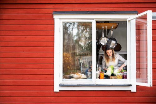 Anastasia Lundqvist står vid ett öppet köksfönster, husets fasad är rött med vitt fönsterbräde. Anastasia håller i olika grönsaker inne i köket.
