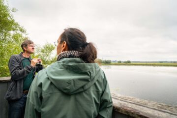 Minh-Xuan och René står uppe på ett fågeltorn, René håller i en kikare och Minh-Xuan ses bakifrån. I bakgrunden syns vatten och kust.